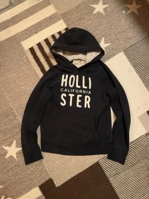 Hollister hoodie - Tja! Säljer denna riktigt goa hollister hoodien med vit ”Hollister California” logga på bröstet och med en riktigt snygg fodrad luva som tillför en cool karaktär till tröjan! Funkar lika bra under kalla vinterdagar som på sena  sommarkvällar och är superenkel att matcha!Tröjan är använd men har inga större defekter. Hör av dig vid minsta fundering! 😁🙌✅
