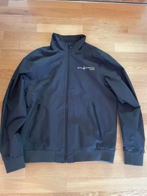 Svart Sail Racing vindjacka Gore-Tex - Snygg svart vindjacka från Sail Racing med hög krage och dragkedja framtill. Jackan har diskret logga på bröstet och Gore-Tex-detalj på ryggen. Tillverkad i slitstarkt material som står emot vind och regn. Perfekt för dig som vill ha en sportig och stilren look.  Fick den i julklapp men ville inte ha den använd max 5 gånger 