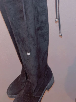 Svarta overknee stövlar i mockaimitation - Säljer ett par svarta overknee stövlar i mjuk mockaimitation. Stövlarna har snörning upptill med silverfärgade detaljer och en rejäl sula för extra komfort. Perfekta för dig som vill ha en trendig och stilren look.