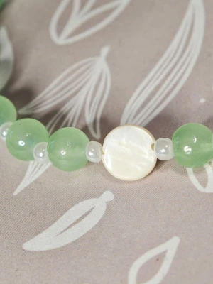 Grönt pärlarmband med snäckdetalj - Stilrent armband med ljusgröna runda glas pärlor och små vita pärlor mellan varje. Armbandet har en platt, rund pärlemor detalj i vitt som ger en unik touch. Perfekt accessoar för att lyfta din outfit med en fräsch och naturlig känsla. Elastiskt armband av hög kvalitet, 18 cm långt.