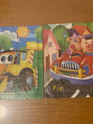 Två färgglada barnpussel - Säljer två roliga barnpussel med tecknade motiv. Ett pussel föreställer en gul grävmaskin med stora ögon under en solig himmel, och det andra visar två glada grisar i en röd bil framför färgglada hus. Perfekt för yngre barn som gillar djur och fordon.