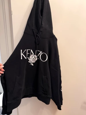 Svart Kenzo hoodie med broderad ros - Svart hoodie från Kenzo med stor vit logga och broderad ros på bröstet. Huva med dragsko och fickor framtill. Kenzo-logga även längs ena ärmen. Oversized passform och mjukt material, perfekt för en chill och trendig look.
