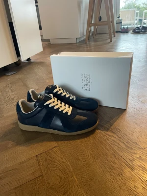 Mörkblå Maison Margielas  - Snygga marinblå Maison Margiela Replica sneakers med ovandel i mocka och läder, vita snören och beige sula. Klassisk low-top siluett som passar perfekt till streetwear och casual outfits. Levereras med originalkartong.