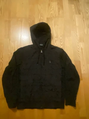 Svart cardigan från Polo Ralph Lauren - Säljer en svart cardigan från Polo Ralph Lauren med dragkedja och broderad logga på bröstet. Jackan har quiltad design, justerbar huva och ribbade muddar. Perfekt för dig som vill ha en stilren och bekväm jacka med exklusiv känsla.