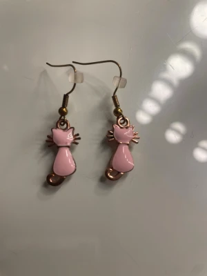 Rosa kattörhängen i metall - Söta örhängen formade som katter i rosa och guldtonad metall. Katterna har en stilren design med tydliga morrhår och krokar i metall. Perfekta för dig som gillar gulliga accessoarer med djurtema.