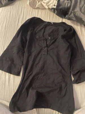 Svart topp med knappar från Vila - En svart topp från Vila med tre knappar framtill och rund halsringning. Toppen har trekvartsärm och är gjord i mjuk bomull, perfekt för en avslappnad och stilren look.