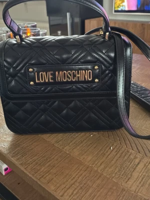 Svart quiltad handväska Love Moschino - Snygg svart quiltad handväska från Love Moschino med guldfärgade detaljer och logotyp framtill. Väskan har både handtag och axelrem, samt ett rymligt huvudfack. Tillverkad i konstläder med elegant rutmönster för en trendig look.