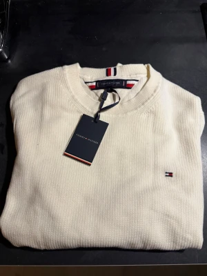Vit stickad tröja Tommy Hilfiger - Klassisk vit stickad tröja från Tommy Hilfiger med rund halsringning och diskret logga broderad på bröstet. Tröjan har ribbade muddar och en stilren look med märkets signaturdetaljer i nacken. Perfekt för en clean och enkel stil i storlek L