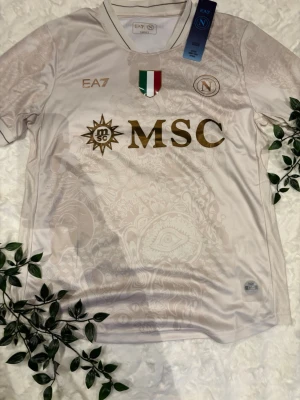 Vit Napoli fotbollströja EA7 - Snygg vit fotbollströja från Napoli med guldfärgade detaljer och MSC-logga på bröstet. Tröjan har ett subtilt mönster i tyget, klubbmärke och italienska flaggan på bröstet. Tillverkad i ett lätt och funktionellt material som passar perfekt till träning eller match.