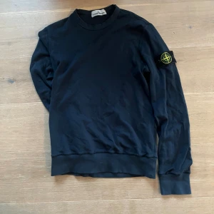 Stone Island tröja - Mörkblå sweatshirt från Stone Island med klassisk rund halsringning och ribbade muddar. Tröjan har den ikoniska Stone Island-loggan på vänster ärm och är tillverkad i mjuk bomull. Perfekt för en avslappnad och stilren look.