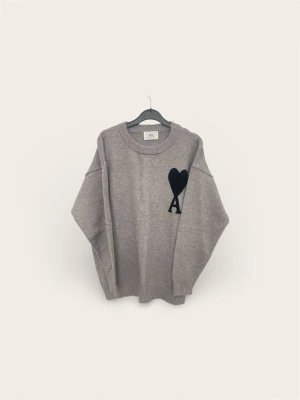 Grå stickad tröja från Ami Paris - Säljer en grå stickad tröja från Ami Paris med svart hjärta och bokstaven A broderat på bröstet. Tröjan har avslappnad passform, rund halsringning och ribbade muddar vid ärmslut och nederkant. Perfekt för dig som gillar stilrena detaljer.