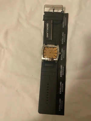 klockor från Excellanc - Säljer stilrena klockor från Excellanc. Den ena har en fyrkantig urtavla i guld och silver, medan den andra har en rund svart urtavla med vita siffror och röda detaljer. Båda har svarta band och metallspänne. Perfekt accessoar för dig som gillar klassisk design. 225kr/ st 