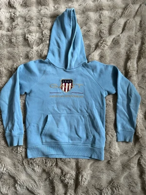 Ljusblå hoodie från GANT - Snygg ljusblå hoodie från GANT med broderad logga och sköld på bröstet. Klassisk känguruficka framtill och huva. Tillverkad i mjuk bomullsblandning, perfekt för en avslappnad och sportig stil. Den har ett superlitet hål som inte syns (se bild 3)