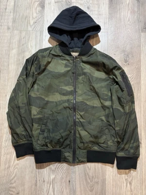 Hollister - Snygg bomberjacka från Hollister i camomönstrad grön nyans. Jackan har svart huva, muddar vid ärmslut och nederkant samt dragkedja framtill. Praktiska fickor och en extra ficka på ärmen. Perfekt för dig som vill ha en streetwear-look.