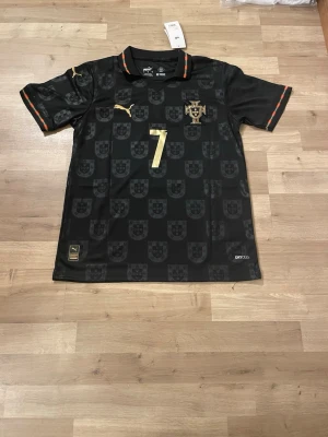 Portugal Ronaldo matchtröja svart Puma - Snygg svart Portugal matchtröja från Puma med guldiga detaljer och nummer 7 på bröstet och ryggen. Ronaldo står tryckt på ryggen. Tröjan har rund hals, korta ärmar och diskret mönster med Portugals sköld. Tillverkad i lätt funktionsmaterial. Storlek S