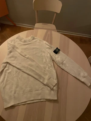 Vit stickad tröja från Stone Island - Säljer en stilren vit stickad tröja från Stone Island med klassisk patch på vänster ärm. Tröjan har rund halsringning, ribbade muddar och nederkant. Perfekt för dig som gillar minimalistisk och clean stil.