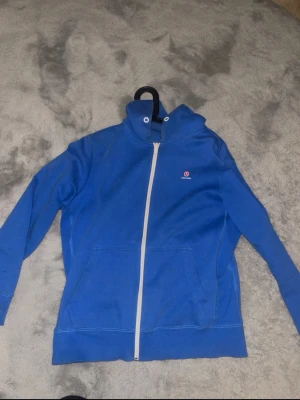 Blå zip hoodie från Björn Borg - Blå hoodie från Björn Borg med vit dragkedja och två stora fickor framtill. Klassisk design med hög krage och diskret logga på bröstet. Ett litet hål på armen men inget man tänker på och tvättas innan! 