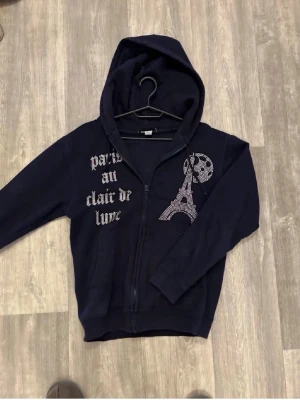 Mörkblå hoodie med Paris-tryck - Snygg mörkblå hoodie med dragkedja och huva. På framsidan finns ett tryck med texten 'paris au clair de lune' och en glittrig Eiffeltorn-design. Hoodien har fickor framtill och är perfekt för dig som gillar streetstyle och coola detaljer.