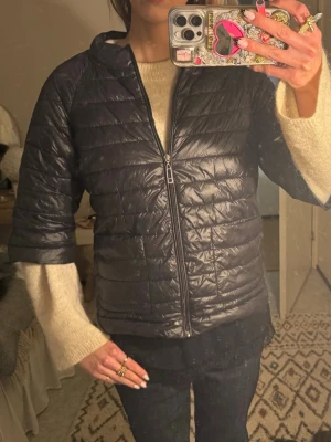 Svart pufferjacka med kort ärm - Snygg svart pufferjacka med korta ärmar och dragkedja framtill. Jackan har en quiltad design och lätt glansigt material som ger en modern look. Perfekt för lager-på-lager och enkel att styla till olika outfits.