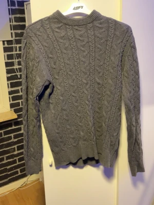 Grå kabelstickad tröja - Säljer en grå kabelstickad tröja med rund hals och långa ärmar. Tröjan har ett klassiskt flätmönster över hela plagget och ribbade muddar vid ärmslut och nederkant. Perfekt för dig som gillar en stilren och mysig look.