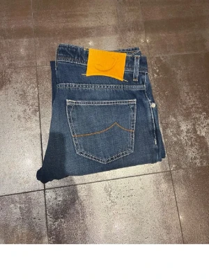 Blå Jacob cohen - Säljer ett par klassiska blå jeans med raka ben och kontrastsömmar. Jeansen har en tydlig gul patch bak i midjan och två bakfickor med dekorativ söm. Perfekta för en avslappnad och stilren look.
