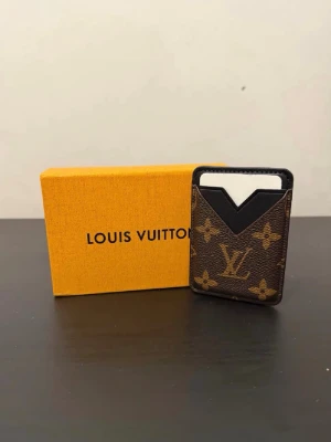  Korthållare - Snygg korthållare i klassiskt brunt monogrammönster. Tillverkad i canvas med svarta läderdetaljer. Slimmad design som passar perfekt för kort och kan fästas (magnet) på baksidan av mobilen. 