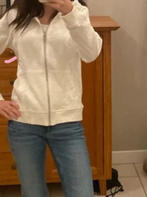 Vit zip hoodie med huva - Säljer en enkel och clean vit hoodie med dragkedja och huva. Hoodien har långa ärmar, ribbade muddar och två fickor framtill. Perfekt för chill dagar eller när du vill ha en basic look. Materialet känns mjukt och bekvämt, troligen bomull eller bomullsblandning.