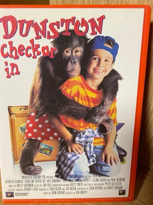 Dunston checkar in VHS - Rolig och färgglad VHS med titeln 'Dunston checkar in'. Omslaget visar en apa och en pojke framför resväskor. Filmen  har ett rött omslag med stora, lekfulla bokstäver och illustrationer. Perfekt för dig som gillar humor och äventyr.