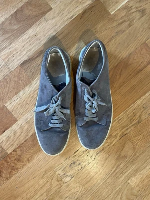 Grå mockasneakers med vita sulor - Grå sneakers i mocka med klassisk låg profil och vita gummisulor. Skorna har rundad tå, platta grå skosnören och enkel design som passar till många olika stilar. Perfekta för dig som gillar en clean och stilren look.