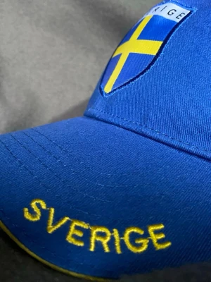 Blå keps med Sverige-flagga Sverigekeps - Sverigekeps, med broderad svensk flagga och texten 'SVERIGE' i gult framtill. Skölden ger en sportig vibe den har en lätt böjd skärm. Perfekt för att visa ditt stöd för Sverige på match eller festival. Eller kanske när du är utomlands .         Jag samlar på kepsar, så de är sparsamt använda och alla har kvar stuket från när de var nya. När jag skickar en keps så gör jag det med mycket vaddering och alltid i en hård kartong. ( är nog ensam om för begagnade kepsar. 