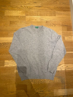 Grå merinoulltröja  - Klassisk grå stickad tröja från United Colors of Benetton, tillverkad i mjuk merinoull. Tröjan har rund halsringning och ribbade muddar vid ärmslut och nederkant. Perfekt för lager på lager och enkel att matcha med olika stilar.