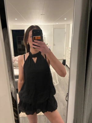 Svart kort klänning med halterneck - Säljer en svart kort klänning med volangkant och halterneck med snygg knutdetalj. Aldrig använt tyvärr och lappen är kvar! Sitter som xs och s! Klänningen. Har även inbyggda shorts vilket är skönt🥰