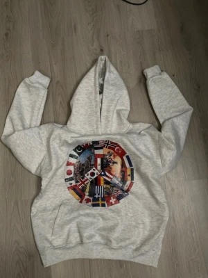 Grå hoodie med flaggor och peace-märke - Snygg ljusgrå hoodie med stort tryck av ett peace-märke på bröstet, dekorerat med olika nationers flaggor och bilder av soldater. Perfekt för dig som gillar unika och statement-plagg. Mjuk och skön med klassisk huva och magficka.