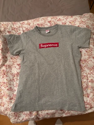 Grå Supreme t-shirt - Säljer en klassisk grå t-shirt från Supreme med det ikoniska röda box logo-trycket på bröstet. T-shirten är tillverkad i 100% bomull och har en normal passform med korta ärmar och rund hals. Perfekt för dig som gillar streetwear och vill ha en clean look.