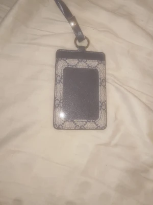 Gucci korthållare med halsband - Snygg korthållare från Gucci i beige och mörkblått med klassiskt GG-mönster. Tillverkad i läder och canvas, har ett fönster för ID eller kort och ett matchande halsband i läder. Perfekt accessoar för att hålla koll på dina kort med stil.