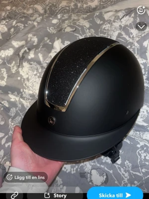 Svart ridhjälm med glitterdetalj - Helt ny oanvänd svart ridhjälm med en glittrig panel upptill och silverfärgade kanter. Hjälmen har justerbart hakband och en klassisk form som passar till ridning. Materialet är matt plast med en dekorativ glittrig yta på ovansidan. Säljer då jag råkade köpa i fel storlek.  Storleken på hjälmen är M son är 55-57 cm. Köpte för 1 500kr kartongen och påsen till hjälmen följer med . Kan enbart frakta om det är genom swish då jag inte har ett bankkonto kopplat här.