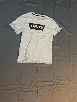 Ljusgrå Levi's t-shirt med logga - Säljer en ljusgrå t-shirt från Levi's med den klassiska svarta loggan tryckt på bröstet. T-shirten har rund halsringning och korta ärmar. Tillverkad i mjuk bomull för en skön känsla och enkel stil.