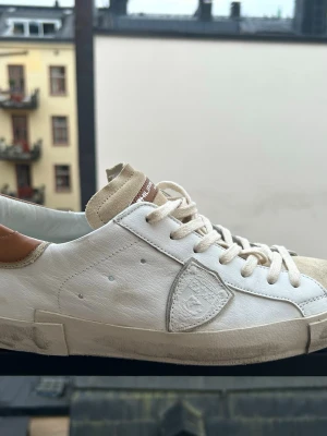 Vita Philippe Model sneakers i läder - Snygga vita sneakers från Philippe Model med beige mockadetaljer på tån och tungan samt bruna detaljer vid hälen. Skorna har klassisk låg profil, vita skosnören och en ikonisk sköldlogga på sidan. Tillverkade i läder och mocka för en clean och stilren look. Endast använt 7 gånger. Storlek 43. Ny pris 380 €. Kan gå ner i pris vid snabb affär. Skicka gärna erbjudande!