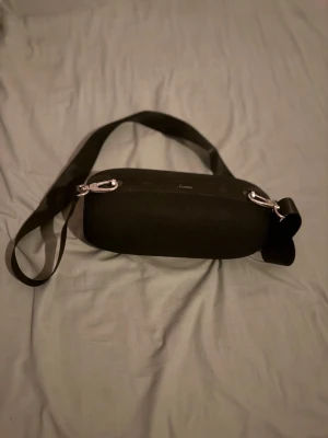 Svart axelväska från Louis Vuitton - Säljer en svart axelväska från Louis Vuitton med minimalistisk design. Väskan har en avlång, rundad form och bred axelrem. Materialet är slitstark textil med silverfärgade metalldetaljer och karbinhakar. Perfekt för dig som gillar stilrena accessoarer.