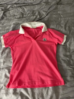 Rosa Moncler piké med vit krage - !!inte äkta!! Snygg rosa pikétröja från Moncler med vit krage och vita ärmslut. Klassisk Moncler-logga på bröstet och broderad text på kragen. Perfekt för dig som vill ha en sportig och trendig look. Passform=xs-s