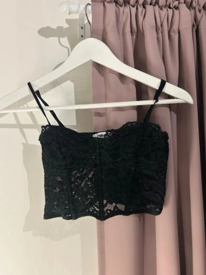 Svart spets topp från New Yorker - Säljer en svart bustier från New Yorker med tunna axelband och vacker spets över hela toppen. Modellen är croppad och har en feminin, figurnära passform. Perfekt att styla till höga jeans eller kjol för en trendig look.