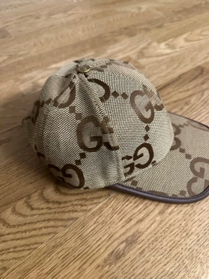 Beige GG-mönstrad keps från Gucci - Snygg beige keps från Gucci med det klassiska GG-mönstret i brunt över hela kepsen. Kepsen har böjd skärm och en metallknapp med Gucci-logga på toppen. Tillverkad i canvasliknande material med exklusiv känsla.