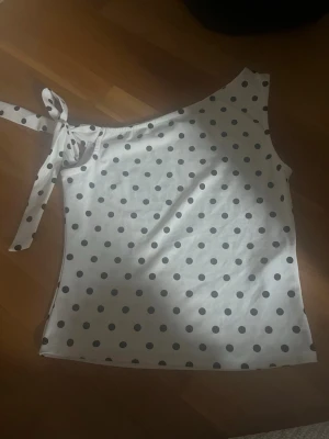 Prickig oneshoulder topp med knyt - Snygg vit oneshoulder topp med svarta prickar och knyt på axeln. Perfekt för dig som vill ha en unik och trendig look. Tunn och mjuk i materialet, passar perfekt till sommaren.