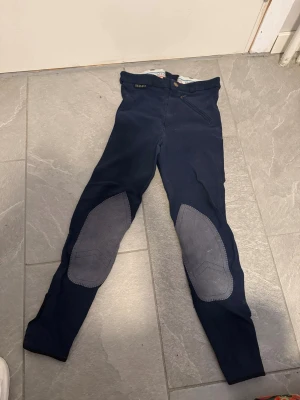 Mörkblå ridbyxor med knäskodd - Säljer ett par mörkblå ridbyxor med gråa knäskodda partier och dragkedja fram. Byxorna har en smal passform och är perfekta för dig som gillar hästar och ridning. Materialet är stretchigt för extra komfort och rörelsefrihet.