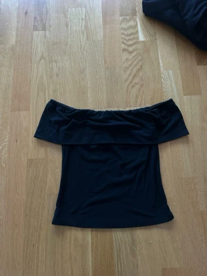 Svart offshoulder topp - Trendig svart offshoulder topp med bred kant upptill som ger en snygg och stilren look. Perfekt att matcha med både jeans och kjol. Tunn och mjuk i materialet, sitter tight och framhäver axlarna.