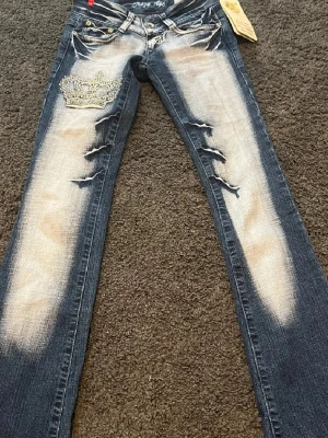 Blå slitna jeans med krona Crazy Age - Säljer ett par blå jeans från Crazy Age . Mått Innerbenet:84cm ytterben:100cm Midjemått: 35,5cm Grenmåttet: 16cm Storlek 32/22. Extremt eftertraktade Crazy Age-jeans med unika detaljer. Extremt lågmidjade, tajta i låren, bootcut och långa – äkta 2000-tals passform.  Väldigt få par finns kvar. Original Crazy Age-lapp kvar.  Jag har sytt ut midjan lite (se sista bilden), men kan sy ihop dem igen innan skick om man vill. Kom privat för mer bilder elr frågor❤️