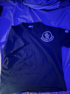 Fet Svart Moncler t-shirt  - Snygg svart t-shirt från Moncler med stor vit logga på bröstet och en liten patch på ärmen. Klassisk rund halsringning och korta ärmar. Perfekt för dig som gillar stilrena märkesplagg.