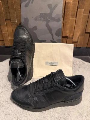 Svarta Valentino rockrunners - Snygga svarta sneakers från Valentino med mattsvarta nitar på hälen och diskret kamouflagemönster. Skornas skick: 8/10! Ny pris för dessa skor ligger runt 6000kr. Skorna är självklart äkta! Storlek 41 men sitter som 42. Skorna har ett fåtal små defekter men inget man ser tydligt. Pris går att diskuteras, hör av er vid frågor eller för flera bilder!🤩 