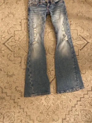 Blå bootcut jeans med slitningar - Säljer ett par ljusblå bootcut jeans med slitningar på framsidan och broderade detaljer i rött på bakfickorna. Jeansen har en tvättad look och vida ben. Perfekta för dig som gillar en avslappnad och trendig stil.