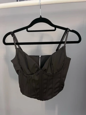 Svart korsettopp med dragkedja - Snygg svart korsettopp med justerbara smala axelband och markerade sömmar. Toppen har en dragkedja i ryggen och croppad passform som ger en edgy look. Perfekt att styla med höga jeans eller kjol.